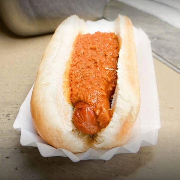 A chili dog in a white bun, served in a paper tray on a wooden surface.