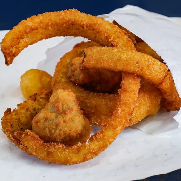 Golden-brown fried onion rings and other fried vegetables spilling out of a white paper bag.