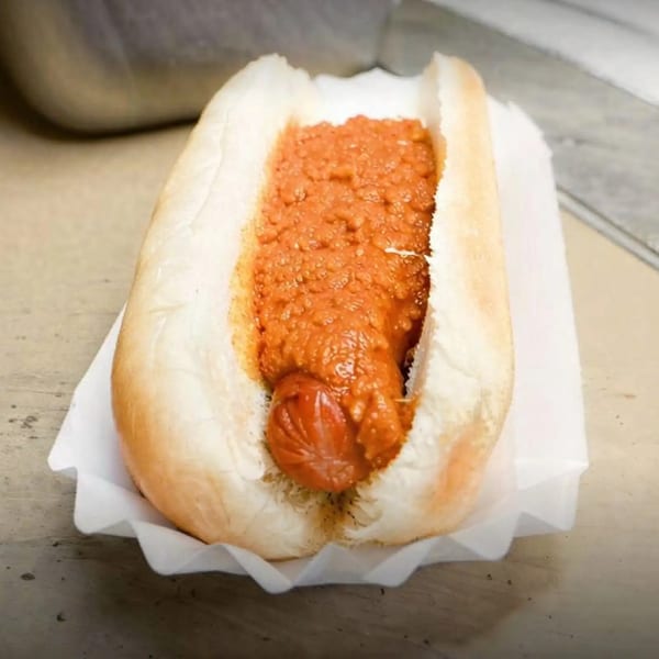 A chili dog in a white bun, overflowing with thick, reddish-brown chili, served in a paper wrapper.